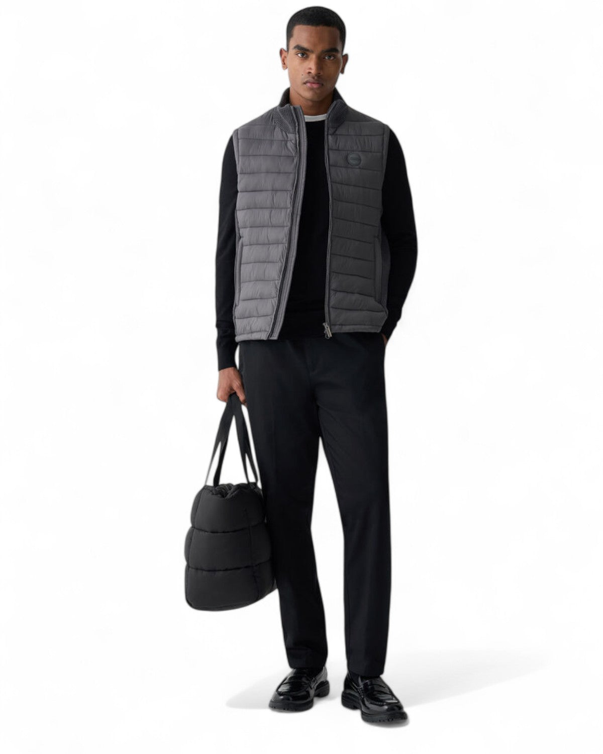 Modello indossa gilet uomo trapuntato con inserti in maglia a costine, abbinato a pantaloni neri e borsa imbottita, look casual invernale.